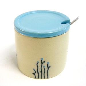 Könnte beinhalten: Eine Zuckerdose aus Keramik mit einem hellblauen Deckel und einem kleinen Löffel. Die Dose hat einen cremefarbenen Boden mit einem schwarzen und blauen Blumendesign.