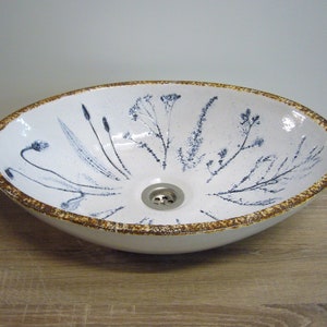 Könnte beinhalten: Weiße ovale Keramikspüle mit braunem Rand und blauem Blumendesign. Die Spüle hat einen Abfluss in der Mitte.