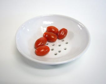 May include: A white ceramic bowl with small holes in the bottom, containing six red cherry tomatoes.