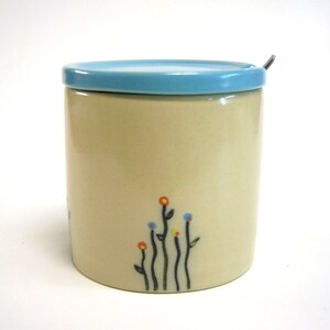 Könnte beinhalten: Eine Keramik-Zuckerdose mit einem hellblauen Deckel und einem Löffel. Die Dose hat einen cremefarbenen Korpus mit einem schwarz-weißen Blumendesign mit orangefarbenen, blauen und gelben Akzenten.