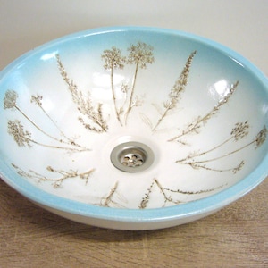 Könnte beinhalten: Ein weißes Keramikwaschbecken mit einem hellblauen Rand und einem braunen Blumendesign. Das Waschbecken ist rund und hat einen Abfluss in der Mitte.