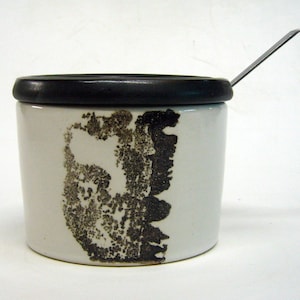 Könnte beinhalten: Eine weiße Zuckerdose aus Keramik mit einem schwarzen Deckel und einem schwarz-weißen abstrakten Design. Die Dose wird mit einem kleinen Löffel aus Metall geliefert.