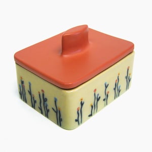 May include: A small ceramic box with a lid. The box is decorated with a pattern of black stems and colorful flowers. The lid is a bright orange color and has a small knob on top.