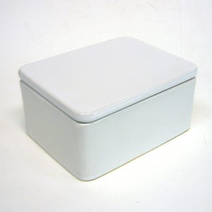 Op de afbeelding: Een rechthoekige, witte keramische doos met bijpassend deksel. De doos heeft afgeronde hoeken en een gladde, glanzende afwerking. Het deksel sluit mooi aan, wat een strak, minimalistisch ontwerp creëert. Ideaal voor het opbergen van kleine items.