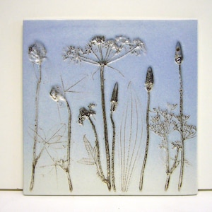 Pode incluir: Um azulejo de cerâmica azul com um desenho estampado de flores e plantas secas. O desenho está em tons de cinza e branco.