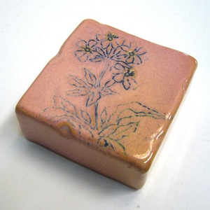 Può includere: Scatola portagioie rettangolare in ceramica con smalto rosa-arancio. La superficie superiore presenta un motivo floreale disegnato a mano in blu scuro. La scatola ha una finitura lucida e una forma leggermente irregolare.