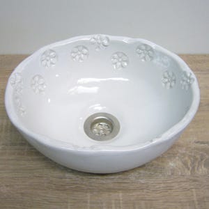 Könnte beinhalten: Ein weißes Keramikwaschbecken mit einem Blumendesign am Rand. Das Waschbecken hat eine glatte, glänzende Oberfläche und einen kreisförmigen Abfluss. Das ovale Waschbecken steht auf einer Holzoberfläche.