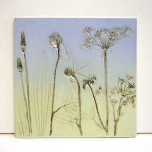 Pode incluir: Um azulejo de cerâmica com um design de flores prensadas. O azulejo apresenta um fundo degradê azul claro e verde com flores prensadas em tons de marrom e branco.