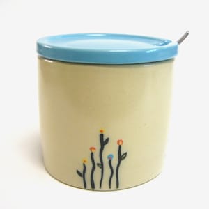 Puede incluir: Un dispensador de azúcar de cerámica con una tapa azul claro y una pequeña cuchara de metal. El dispensador tiene un cuerpo de color crema con un diseño de flores negras y coloridas.