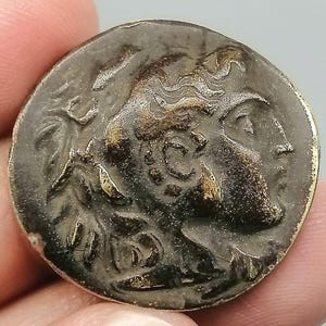 May include: A bronze coin featuring a profile of a person with detailed hair and facial features. The coin has a dark, aged appearance with visible wear and a golden rim. The coin is held in a hand.