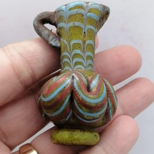 May include: A small, antique glass vessel with a handle. The vessel features a bulbous base and a flared top, adorned with colorful stripes of red, blue, and white against a yellow-green background. The handle is a dark brown color.