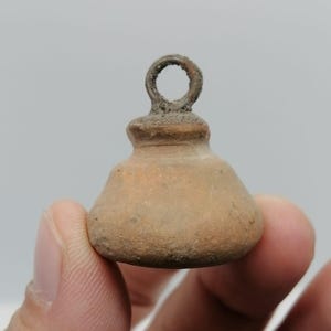 Pode incluir: Um pequeno objeto em forma de sino com uma argola no topo. O objeto é de cor castanho claro com uma superfície texturizada. A argola é de um castanho enferrujado mais escuro. O objeto é segurado entre dois dedos.
