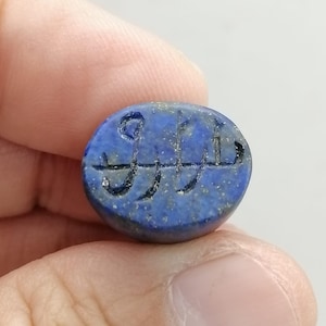 May include: Oval-shaped, deep blue lapis lazuli stone with etched markings. The stone is held between fingers and thumb. The markings appear to be an ancient script or symbol. The stone has a polished surface and a slightly irregular shape.