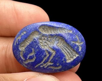 May include: A deep blue, oval-shaped stone with a carved design of a bird-like creature. The carving is in a lighter color, creating contrast against the dark blue background. The stone is held between fingers against a black background.
