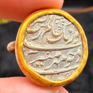 May include: A round, antique coin with gold-colored edging. The coin's center features intricate silver-colored script. The coin is held between fingers, suggesting its small size and historical significance. The coin's design is detailed and well-preserved.