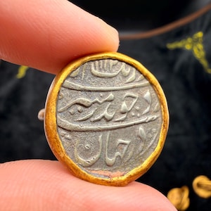 Puede incluir: Una moneda redonda y antigua con borde dorado. El centro de la moneda presenta texto grabado descolorido en una escritura ornamentada. La moneda se sostiene entre dos dedos, lo que sugiere su pequeño tamaño y su importancia histórica.