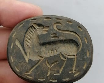 May include: Oval, dark brown carved wooden pendant featuring a stylized animal, possibly a horse or zebra. The animal is depicted in profile with detailed lines and a textured surface. The pendant has a decorative border.