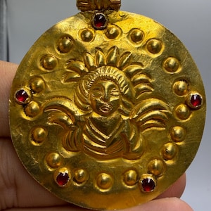May include: A circular gold pendant featuring a central embossed figure with wings and a radiating halo. The surface is decorated with small raised gold spheres and five inset deep red gemstones arranged in a circular pattern around the figure.