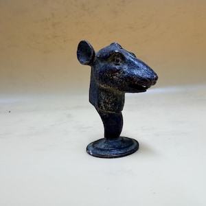 Pode incluir: Escultura em bronze com cabeça de rato, com rosto detalhado, orelhas e base arredondada. A escultura tem uma pátina escura, sugerindo idade ou um acabamento envelhecido. A cabeça do rato está em pé.