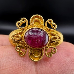 May include: A gold ring featuring an oval, deep red gemstone. The ring has an ornate, filigree design with swirling patterns around the gemstone. The ring is set against a dark background.