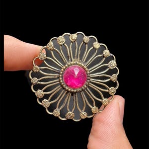 May include: A vintage-style brooch with a central, faceted pink gemstone. The brooch features a detailed, openwork design in a gold tone, with radiating lines and small floral accents around the edge. The brooch is held between two fingers against a black background.