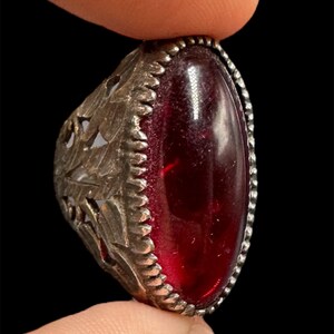 May include: A silver-toned ring featuring an oval, deep red gemstone. The ring's band has an intricate, openwork design with leaf-like patterns. The gemstone is set within a beaded border, creating a vintage aesthetic.