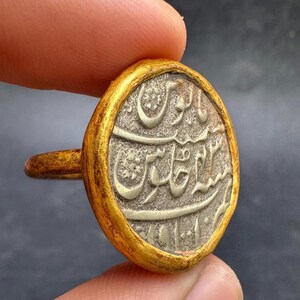 May include: A gold-toned ring featuring a circular, silver-colored coin with intricate script. The coin is set within a gold bezel and attached to a simple band. The ring is held between fingers, showcasing its antique appearance.