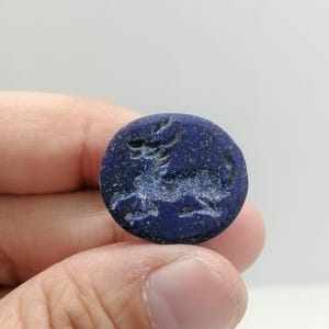 May include: A round, dark blue stone with a carved image of an animal. The stone is held between fingers and thumb. The animal carving is light blue and appears to be a lion or similar feline.