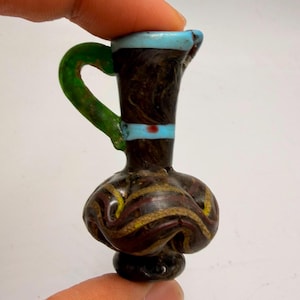 Puede incluir: Una jarra de vidrio en miniatura con cuerpo marrón oscuro y patrones en espiral de color amarillo y rojo oscuro. El recipiente tiene un asa verde brillante, un borde azul claro y una banda azul claro alrededor del cuello con un punto rojo.