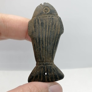 May include: A dark, weathered fish-shaped pendant. The pendant features a detailed design with vertical lines and a small hole near the tail. The fish has a circular eye and a textured surface, possibly an antique or vintage item.