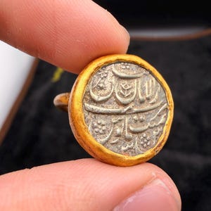 Puede incluir: Una moneda redonda y antigua con borde dorado. El centro de la moneda presenta textos y diseños grabados intrincados en plata. La moneda se sostiene entre dos dedos sobre un fondo oscuro, resaltando sus detalles.