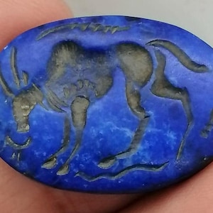 Op de afbeelding: Ovale, diepblauwe edelsteen met een gesneden ontwerp van een dier, mogelijk een stier of koe. Het dier is afgebeeld in een gestileerde, oude stijl, met details geëtst in een contrasterende kleur. Het oppervlak van de steen heeft een natuurlijk, getextureerd uiterlijk.