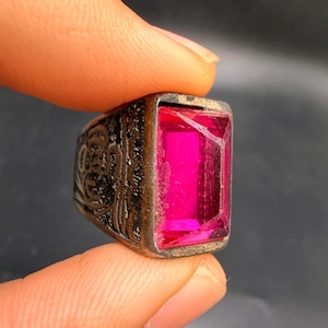 Puede incluir: Un anillo plateado con una gema rectangular grande de color rosa brillante. La banda del anillo presenta un diseño grabado y ornamentado. La gema está engastada en un bisel. El anillo está sujeto por un dedo.