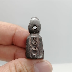 Könnte beinhalten: Ein kleiner, dunkel gefärbter Anhänger mit einer Schlaufe oben und einem geschnitzten Design auf der Vorderseite. Das Design scheint eine stilisierte Figur oder ein Symbol zu sein. Der Anhänger wird in der Hand einer Person gehalten.