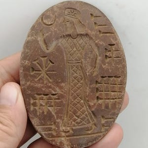 May include: Oval-shaped, brown stone carving featuring a detailed figure in a long, patterned robe and headdress. The carving includes symbols and a crescent moon. The stone is held in a hand.