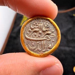 May include: A round, antique coin with Arabic script. The coin has a silver center and a gold-colored rim. The coin is held between two fingers, with a dark background.