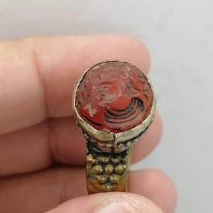 May include: An antique ring with a red stone and a carved design. The ring has a gold-coloured band with a textured pattern. The stone is set in a silver-coloured bezel.