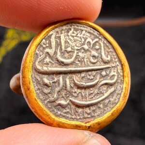 May include: A round, antique coin with intricate Arabic script and a gold-colored border. The coin's silver center shows signs of wear, suggesting age and historical significance. The coin is held between two fingers.