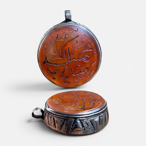 Op de afbeelding: Twee antieke zilveren hangers met een cirkelvormige, oranjebruine steen. De steen is gegraveerd met Arabisch schrift. De hangers hebben een zilveren lus om op te hangen en een decoratieve zilveren rand.