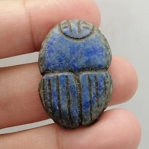 Op de afbeelding: Een blauwe scarabee keversnijwerk met gedetailleerde lijnen en een getextureerd oppervlak. De kever is diepblauw met donkere lijnen en wordt in een hand gehouden. De snijwerk is waarschijnlijk gemaakt van steen.