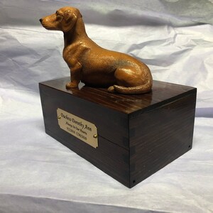 Puede incluir: Una figura de perro salchicha marr&oacute;n se encuentra sobre una caja de madera oscura. La caja tiene una placa dorada que dice "Duchess Dorecky Ann, Always In Our Hearts, 9/12/2018 - 7/08/2020". La figura es una representaci&oacute;n detallada del perro.