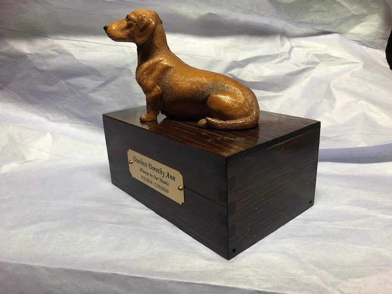 Puede incluir: Una figura de perro salchicha marr&oacute;n se encuentra sobre una caja de madera oscura. La caja tiene una placa dorada que dice "Duchess Dorecky Ann, Always In Our Hearts, 9/12/2018 - 7/08/2020". La figura es una representaci&oacute;n detallada del perro.
