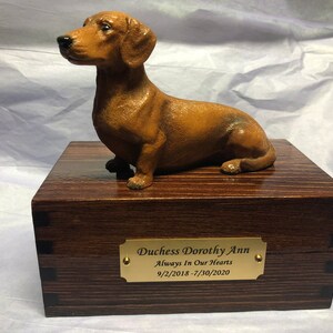 Puede incluir: Una figura de perro salchicha marr&oacute;n se encuentra sobre una caja de madera con una placa de lat&oacute;n. La placa dice "Duchess Dorothy Ann Always In Our Hearts 9/2/2018 - 7/30/2020". La caja tiene un acabado de vetas de madera oscura y est&aacute; construida con uniones de cola de milano.