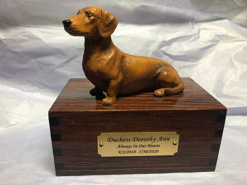Puede incluir: Una figura de perro salchicha marr&oacute;n se encuentra sobre una caja de madera con una placa de lat&oacute;n. La placa dice "Duchess Dorothy Ann Always In Our Hearts 9/2/2018 - 7/30/2020". La caja tiene un acabado de vetas de madera oscura y est&aacute; construida con uniones de cola de milano.