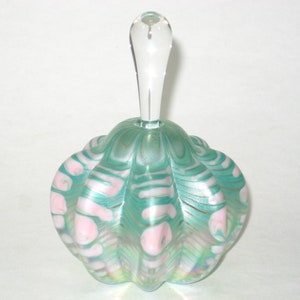May include: A decorative glass perfume bottle with a clear, elongated stopper. The bottle has a rounded, scalloped shape with a teal and pink swirl pattern. The glass has an iridescent sheen.