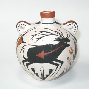 May include: A hand-painted ceramic flask with a cream-colored base. The flask features a black deer design with an orange arrow, black antlers, and decorative elements. The rim and handle accents are brown.