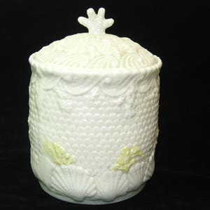 May include: Cream-colored ceramic jar with lid, featuring a textured surface with a scale-like pattern. The jar is adorned with seashell and coral designs, and has a decorative finial on the lid. The jar is approximately 6 inches tall.
