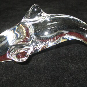 Op de afbeelding: Een heldere glazen dolfijnsculptuur met een gebogen lichaam en een puntige rugvin. De sculptuur heeft een glad, gepolijst oppervlak en lijkt van kristal te zijn gemaakt. De naam "Tzora" is aan de zijkant ge&euml;tst.