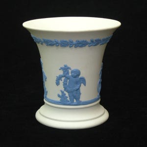 May include: A white ceramic vase with a blue and white design. The vase has a wide mouth and a narrow base. It features a relief design of a cherub holding a bunch of grapes.