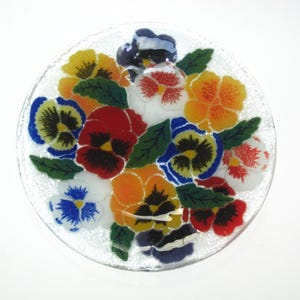 May include: A clear, round glass plate decorated with a colorful floral design. The design features a bouquet of pansies in shades of yellow, orange, red, blue, and white, with green leaves. The plate is on a white background.